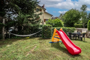 Un parque infantil con un tobogán rojo y una estructura de escalada amarilla, rodeado de césped y árboles, con un edificio al fondo.