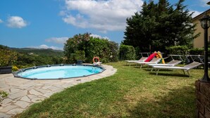 A swimming pool with a stone deck, surrounded by grass and trees, with a view of hills in the background.