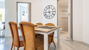 A dining area with wicker chairs, a wooden table, a wall clock, and a door leading to a bathroom. - Holiday Apartment "Casa Feliz" with Shared Pool, Private Terrace & Wi-Fi (Mijas)