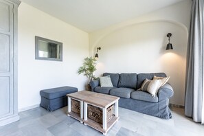 Una sala de estar con un sofá gris, una mesa de centro de madera, un banco, una planta pequeña y una lámpara de pared.