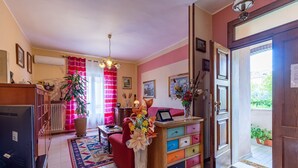 Ein Wohnzimmer mit einem roten Sofa, einer Holzkassette mit bunten Schubladen, einer großen Vase mit Blumen und einer Tür, die zu einem Außenbereich führt.
