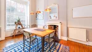 Un coin repas avec une table en bois, des chaises et un tapis à motifs bleus. On y trouve une cheminée avec un mur en pierre, une fenêtre donnant sur des bâtiments et un radiateur fixé au mur.