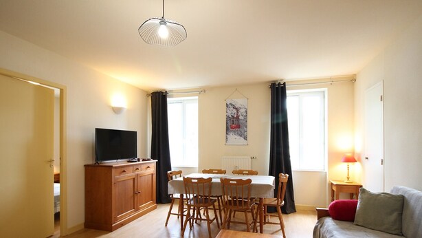 A living room with a wooden table, a sofa, a TV on a cabinet, and a dining area with chairs and a table.