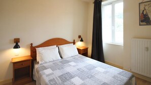 A bedroom with a wooden headboard, two bedside tables with lamps, a quilted bedspread, a window with a curtain, and a poster on the wall.