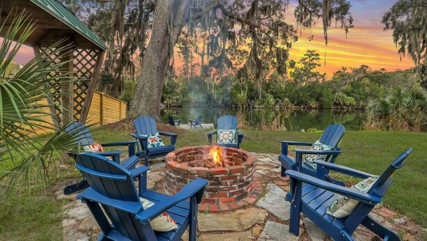 A fire pit surrounded by four Adirondack chairs with cushions, set on a stone patio with a view of a lake and trees at sunset.
