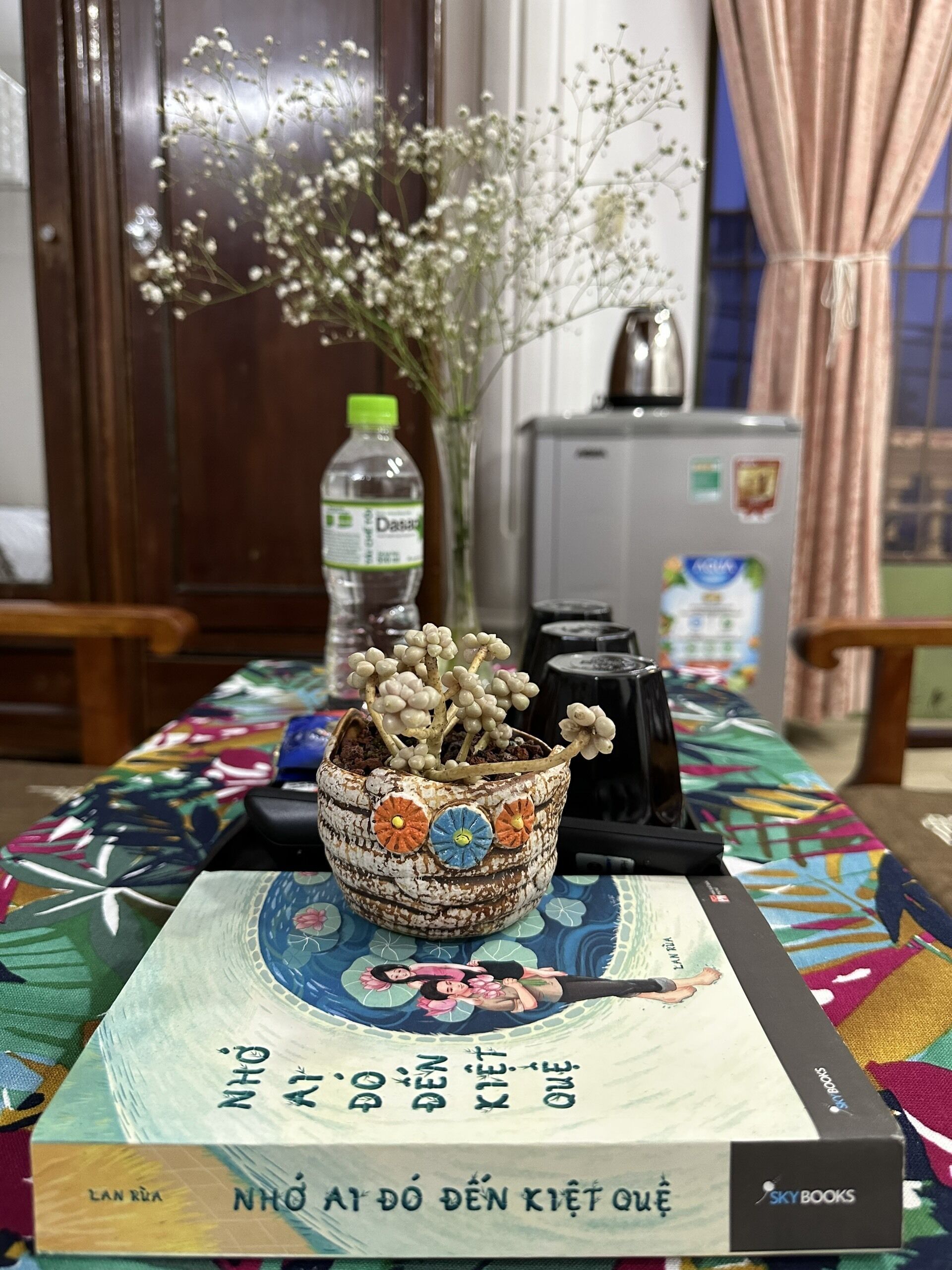 Une table avec un livre intitulé « Nhớ về ngày điên khiết quy » et un pot décoratif contenant des fleurs séchées.
