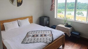 Un dormitorio con una cama de madera, una mesita con un florero, un refrigerador y una ventana con vista a un lago.