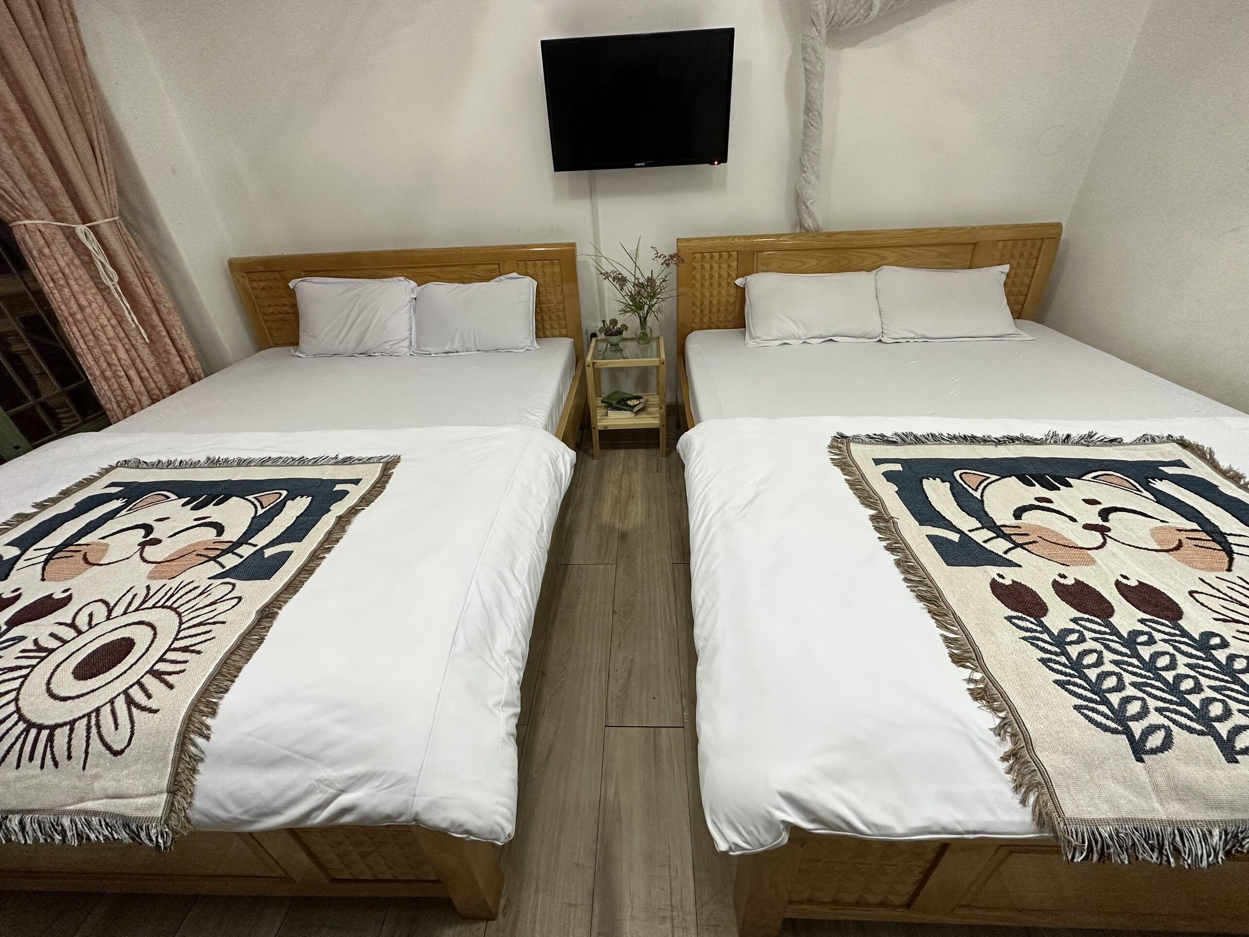 A double bed room with two beds, each with a white bedspread and a decorative blanket. There is a small bedside table with a vase of flowers between the beds.