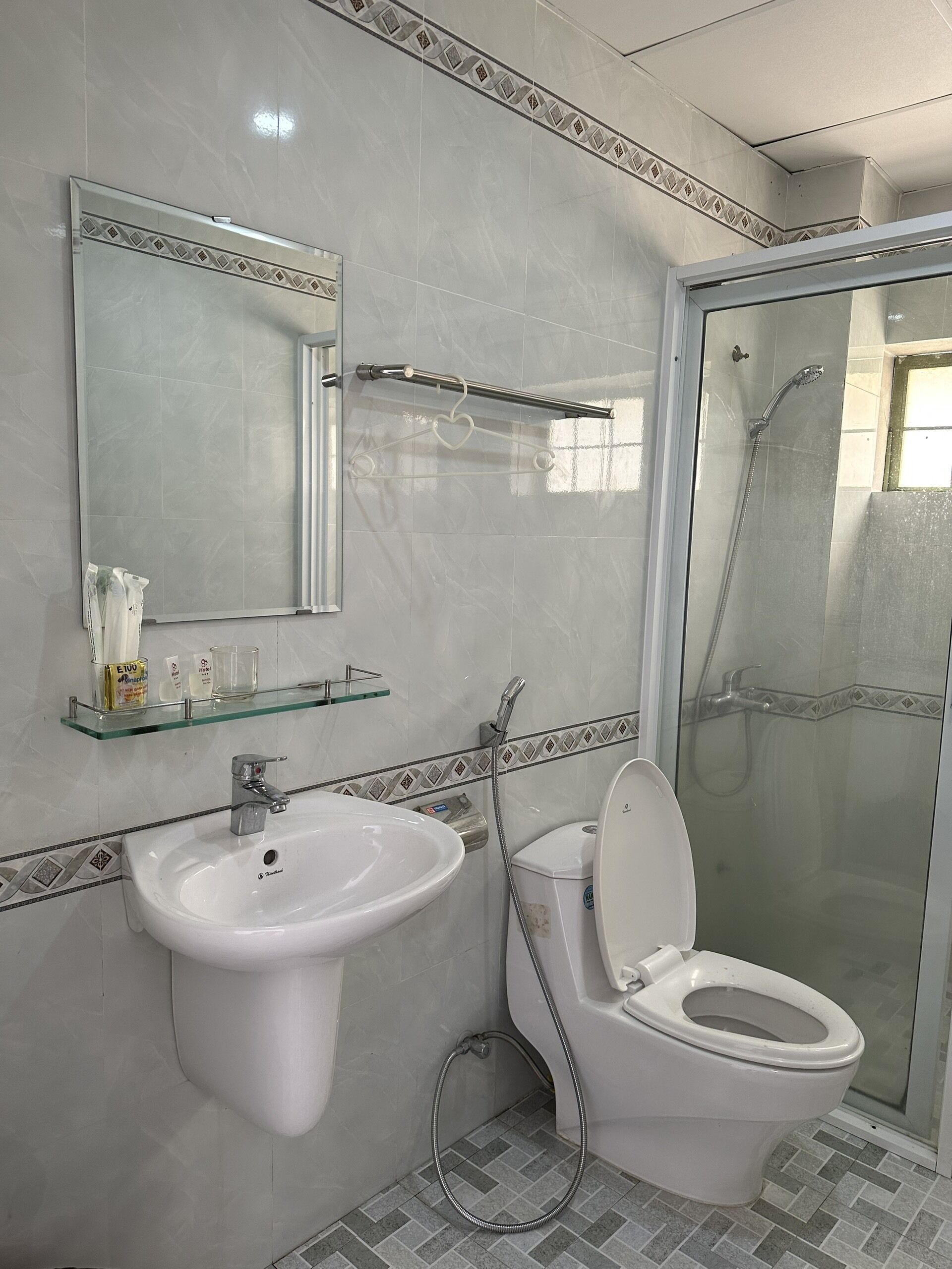 A bathroom with a white sink, toilet, and shower. There is a glass shelf above the sink with toiletries.