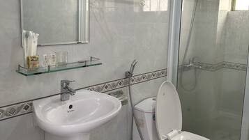 A bathroom with a white sink, toilet, and shower. There is a glass shelf above the sink with toiletries.