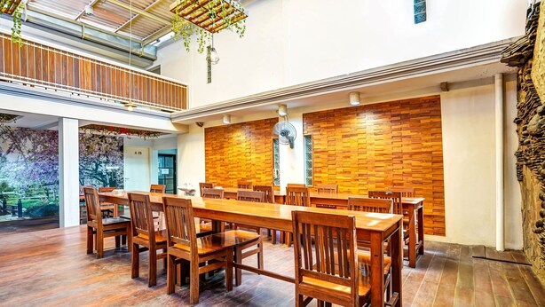 A spacious dining area with wooden tables and chairs, a brick wall, and a view of a garden through large windows. - OYO Dupak Inn (Surabaya)