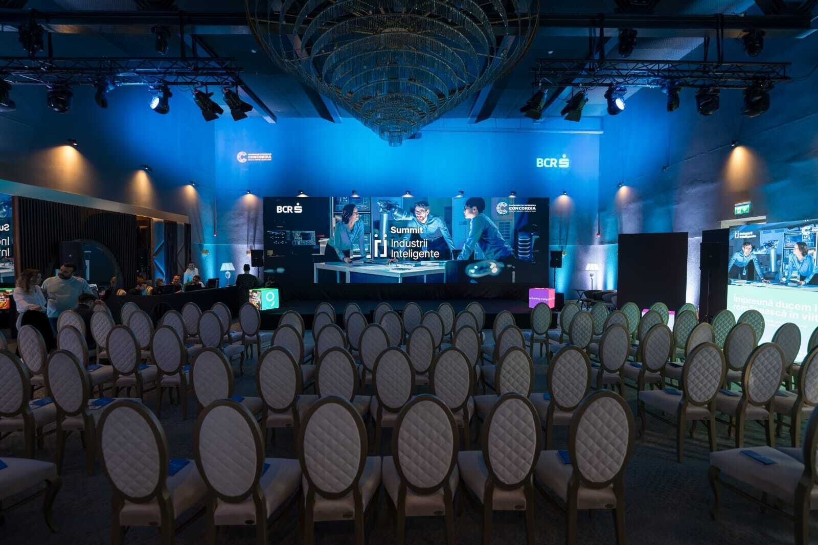 Sala de conferência com fileiras de cadeiras voltadas para um palco com uma tela grande exibindo uma apresentação.