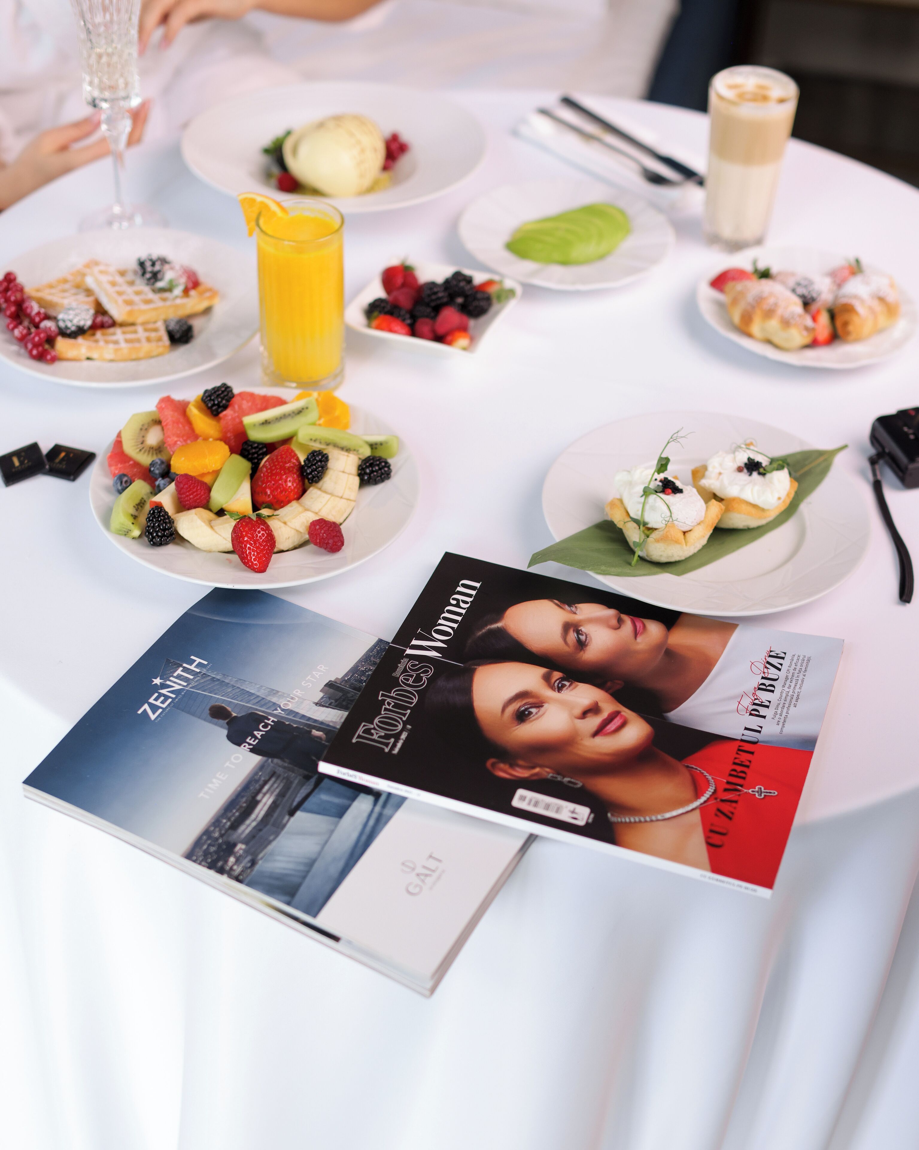 Een tafel met diverse gerechten, waaronder fruit en gebak, en een tijdschrift met de titel 'Forbes Woman'.
