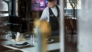 Interior de un restaurante con un camarero con chaleco, una mesa de comedor dispuesta con platos, vasos y un holder para servilletas.