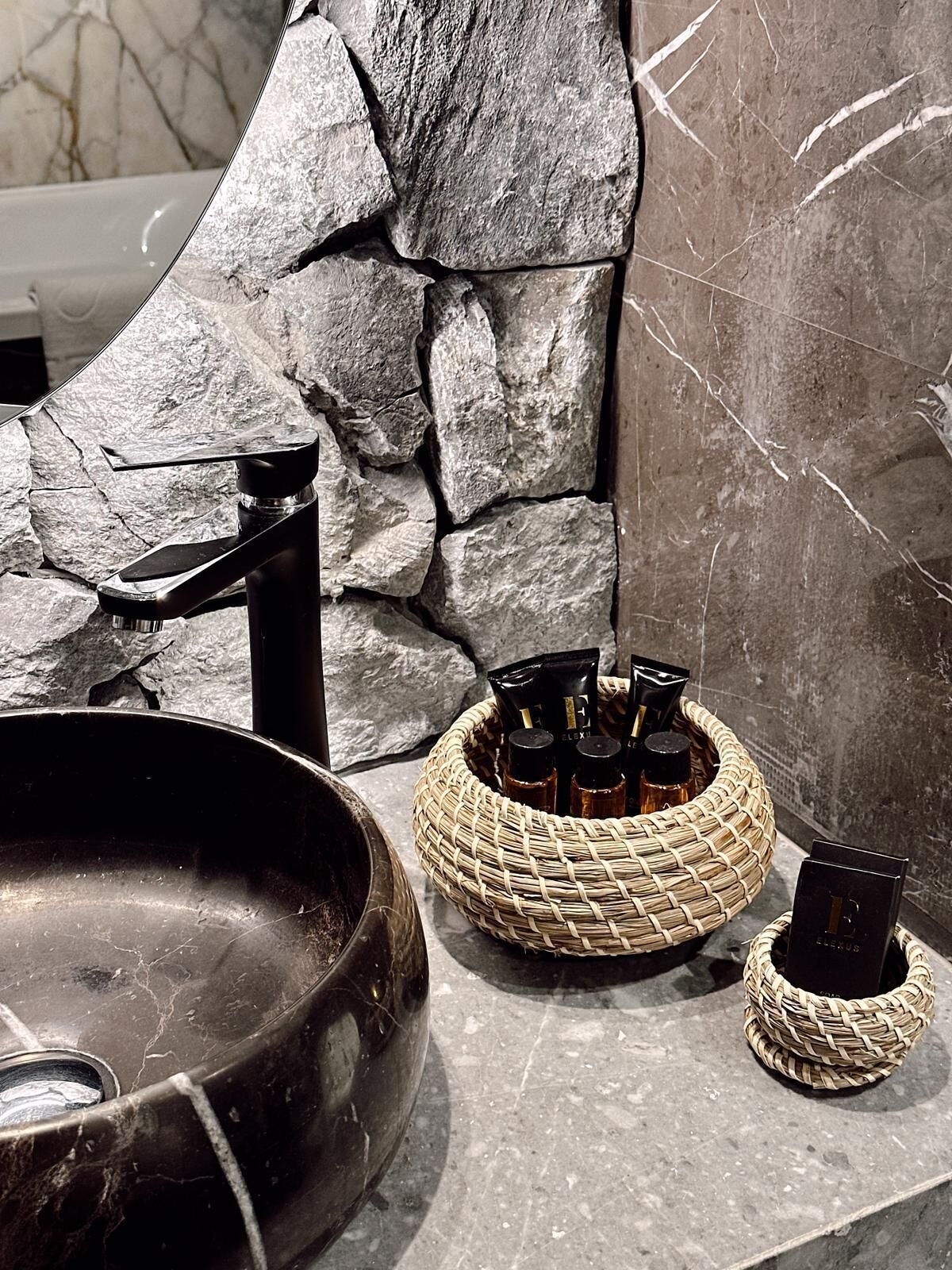 A bathroom with a stone wall, a black sink, and a woven basket with toiletries.