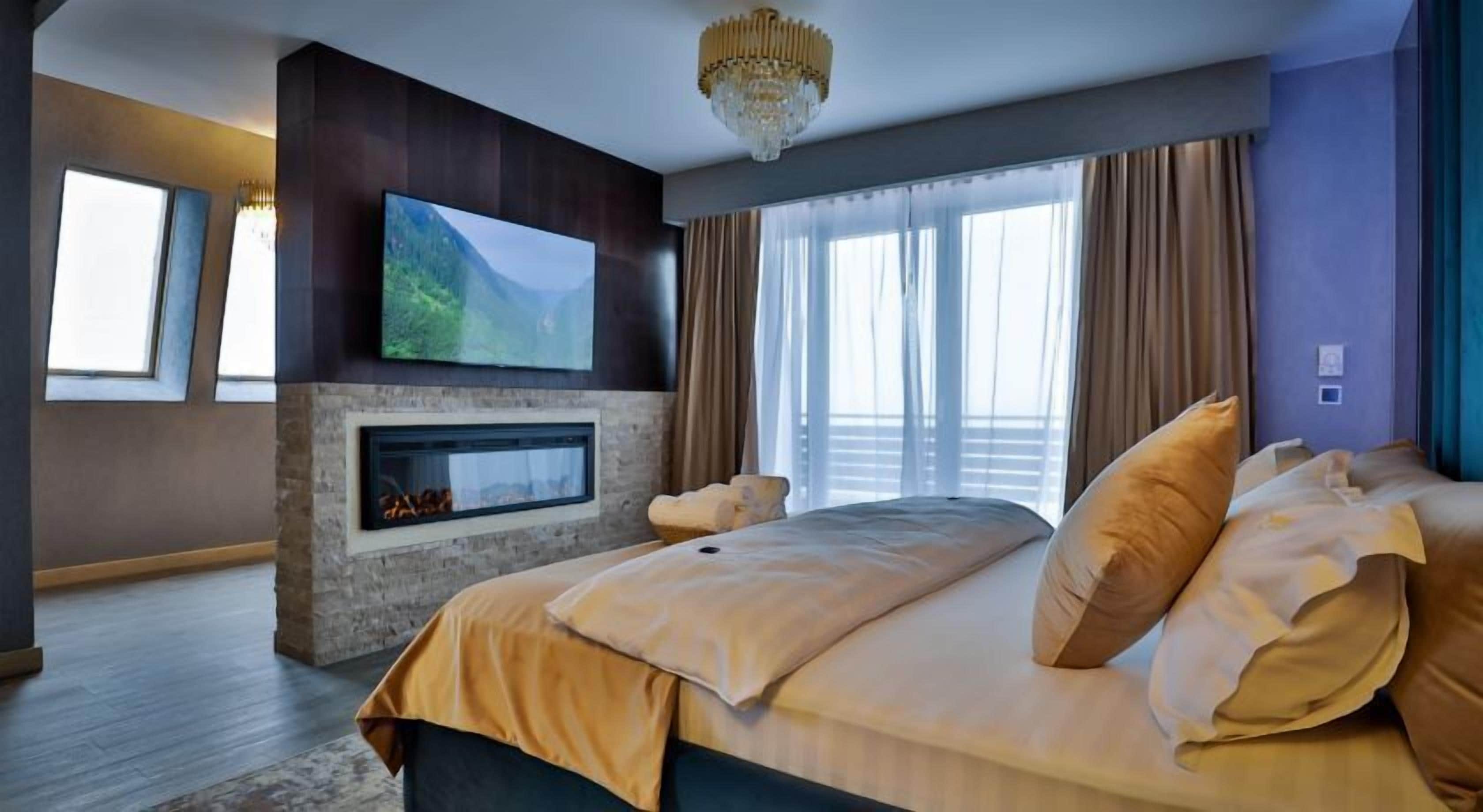 A modern hotel room with a large bed, a flat-screen TV mounted on the wall, a fireplace, and large windows with curtains.