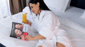 Una persona en un albornoz blanco recostada sobre una cama, con una revista, un vaso de zumo de naranja y una bandeja de desayuno que incluye gofres, frutos rojos y un postre sobre una mesa blanca.