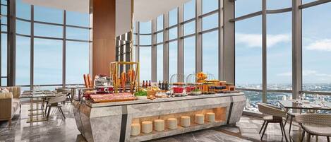 Habitación de hotel en un edificio alto con un ventanal amplio que ofrece vistas a la ciudad, un mostrador de recepción de mármol y muebles modernos.