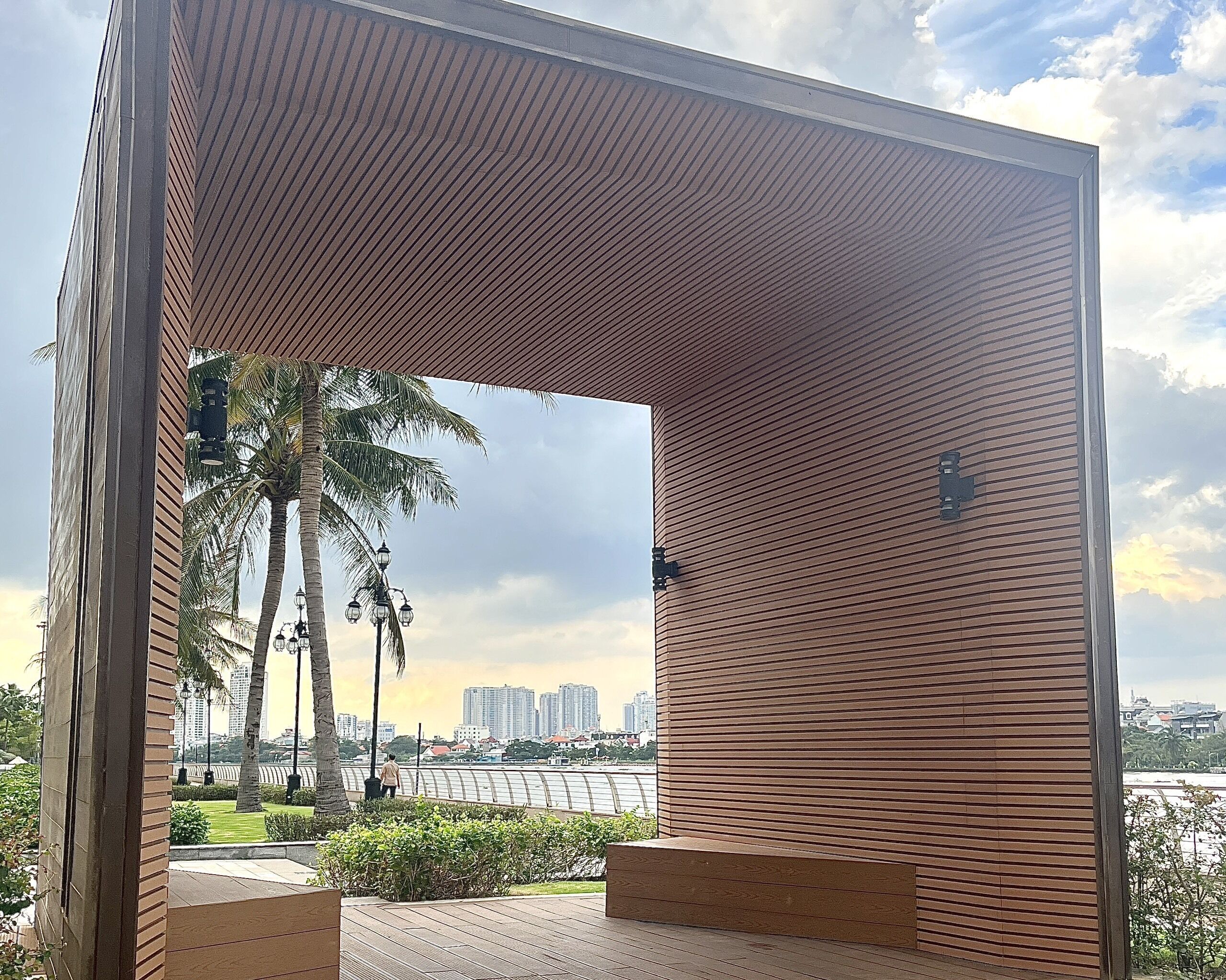 Un pabellón moderno con techo de listones, rodeado de jardines con palmeras y vistas a un río y al horizonte de la ciudad.