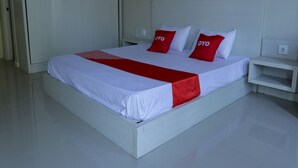 Una cama bien hecha con ropa blanca y detalles en rojo, una mesita de noche blanca a un lado y una puerta cerrada al fondo.