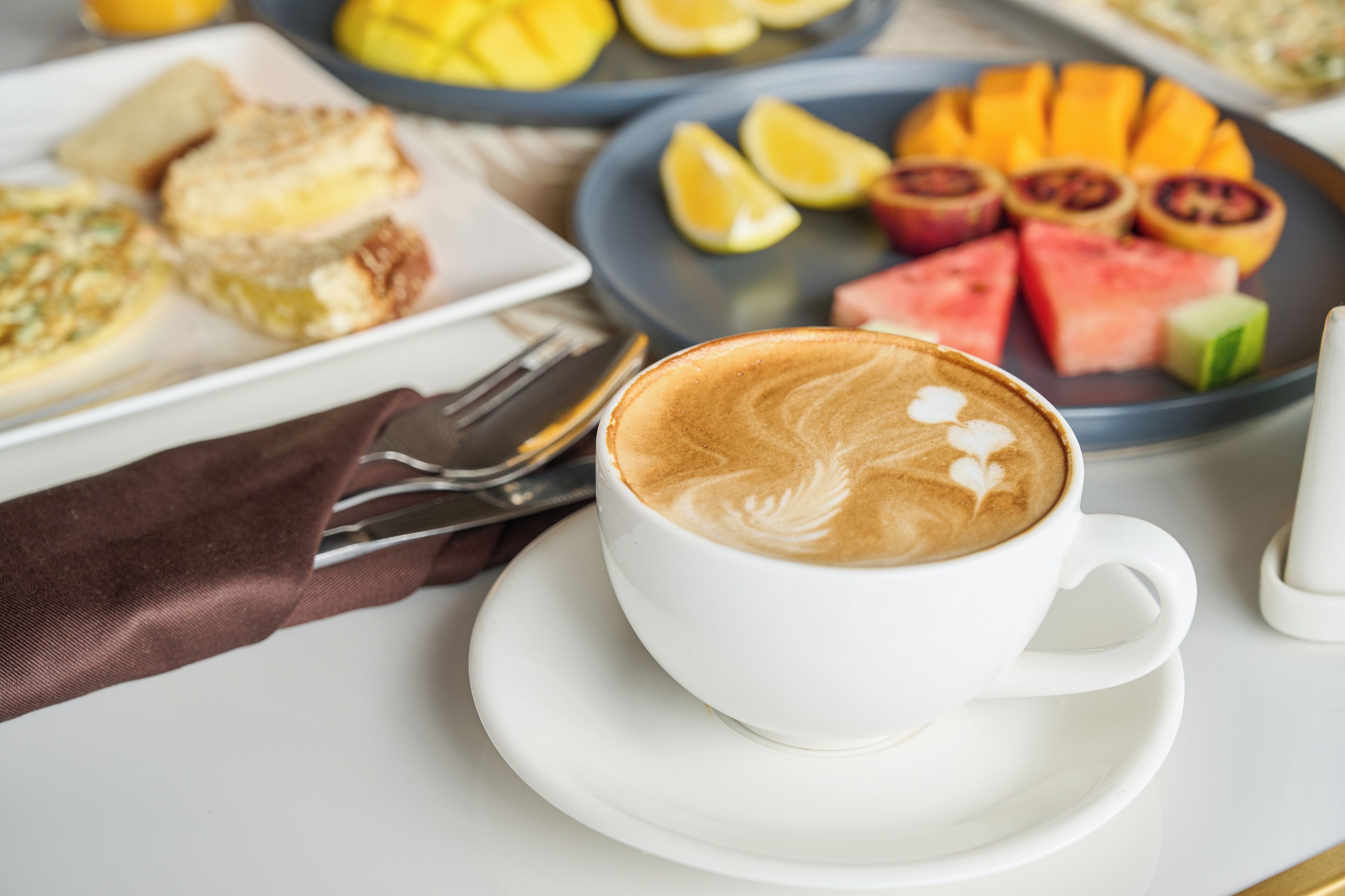 A cup of coffee with latte art on a saucer, a plate with sliced fruits, and a plate with pastries.