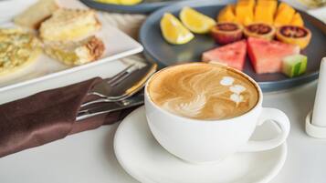 Une tasse de café décorée avec un motif en latte, posée sur une soucoupe, une assiette de fruits coupés et une assiette de pâtisseries.