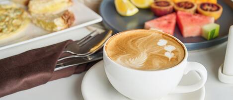 A cup of coffee with latte art on a saucer, a plate with sliced fruits, and a plate with pastries.