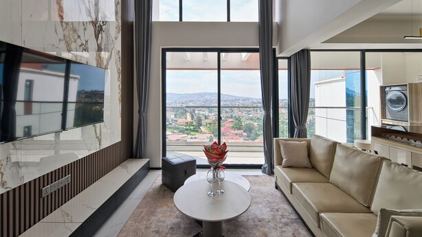 A modern living room with a marble wall, a round table with a vase, a beige sofa, and a large window offering a city view. - Oasis Park (Kigali)