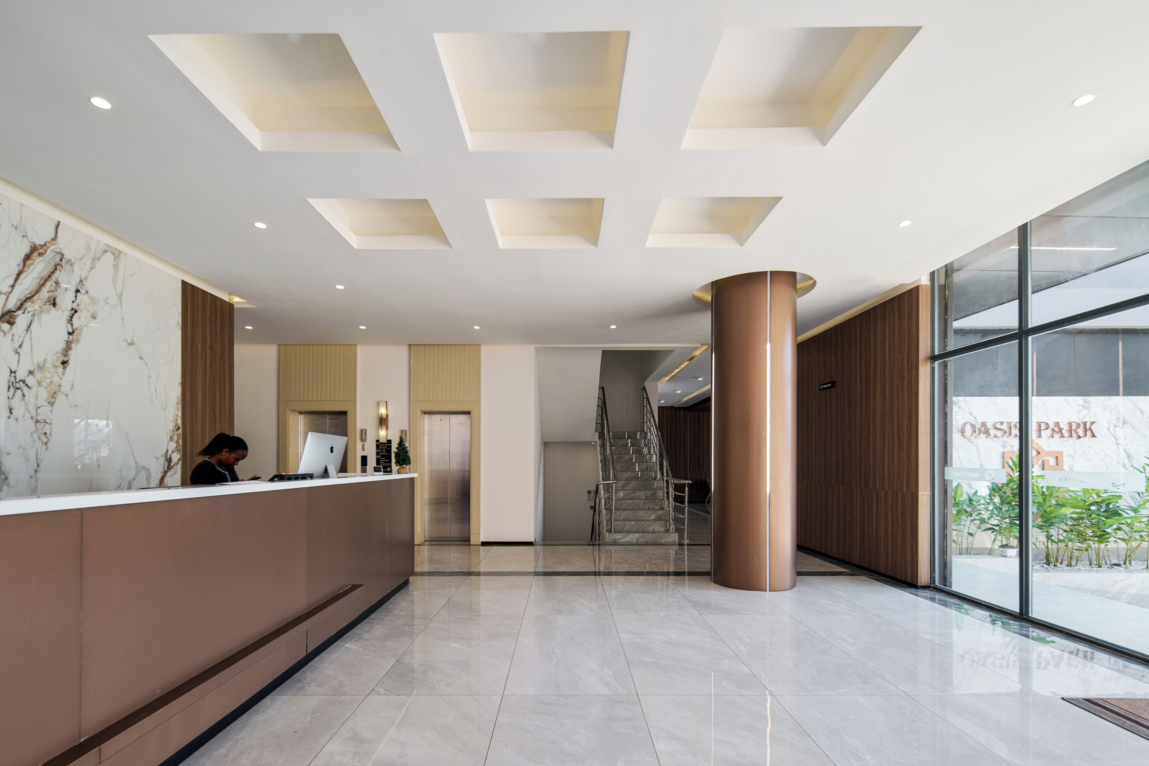 A modern hotel lobby with a reception desk, a staircase, and a view of the outside.