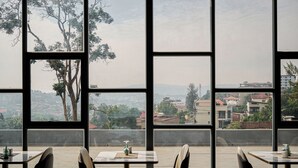 A restaurant with large windows offering a view of a cityscape. Tables are arranged with chairs, and a vase is visible on one table. - Oasis Park (Kigali)