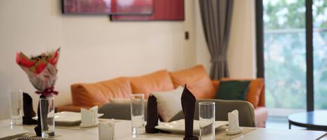 A dining area with a table set for two, a sofa in the background, and a window with a view.