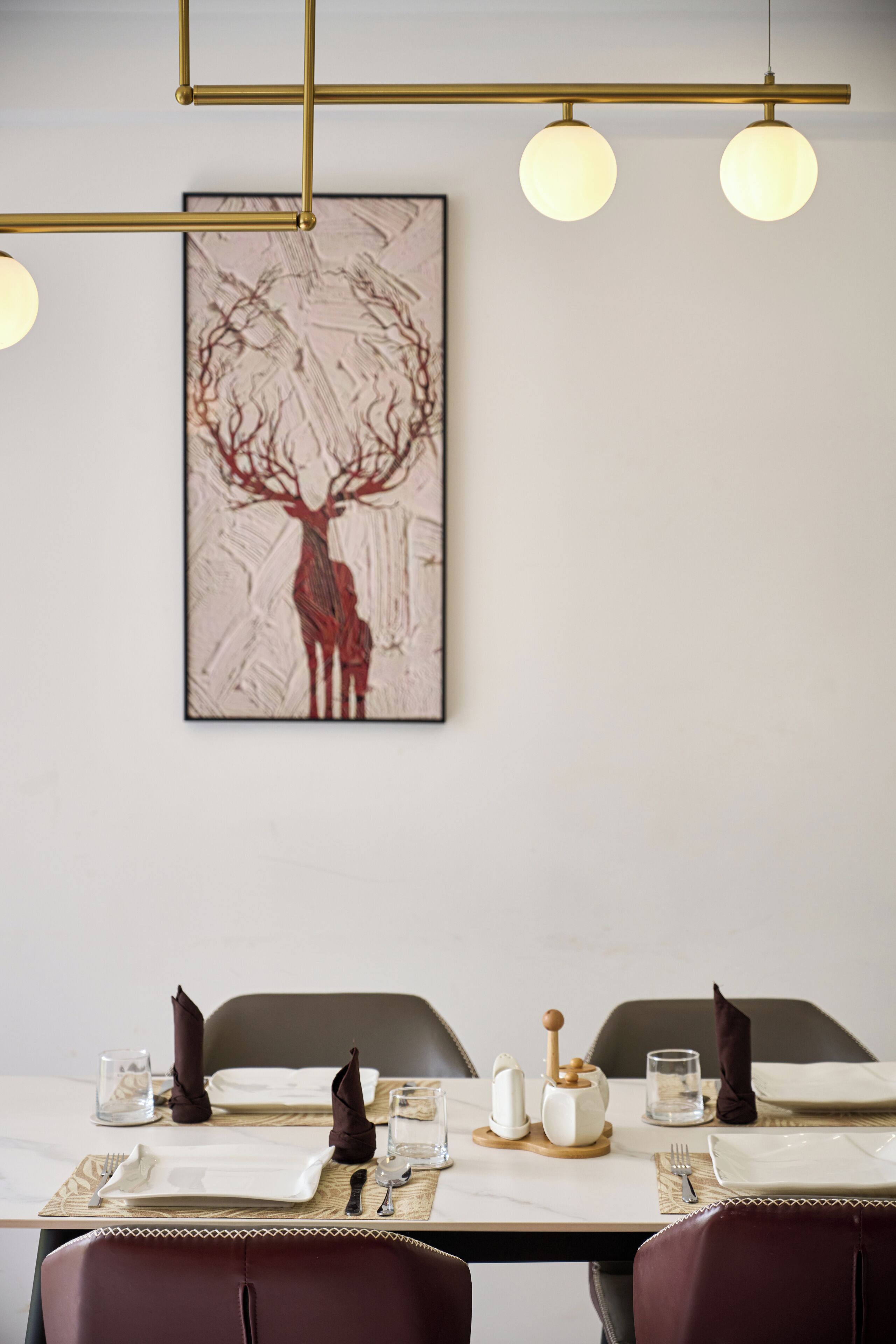A dining area with a table set for two, a framed artwork of a deer, and a modern light fixture above.