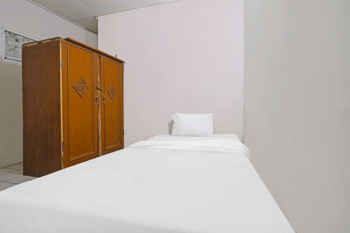 Een enkel bed met een witte matras en kussen, een houten garderobe met twee deuren en een effen witte muur.