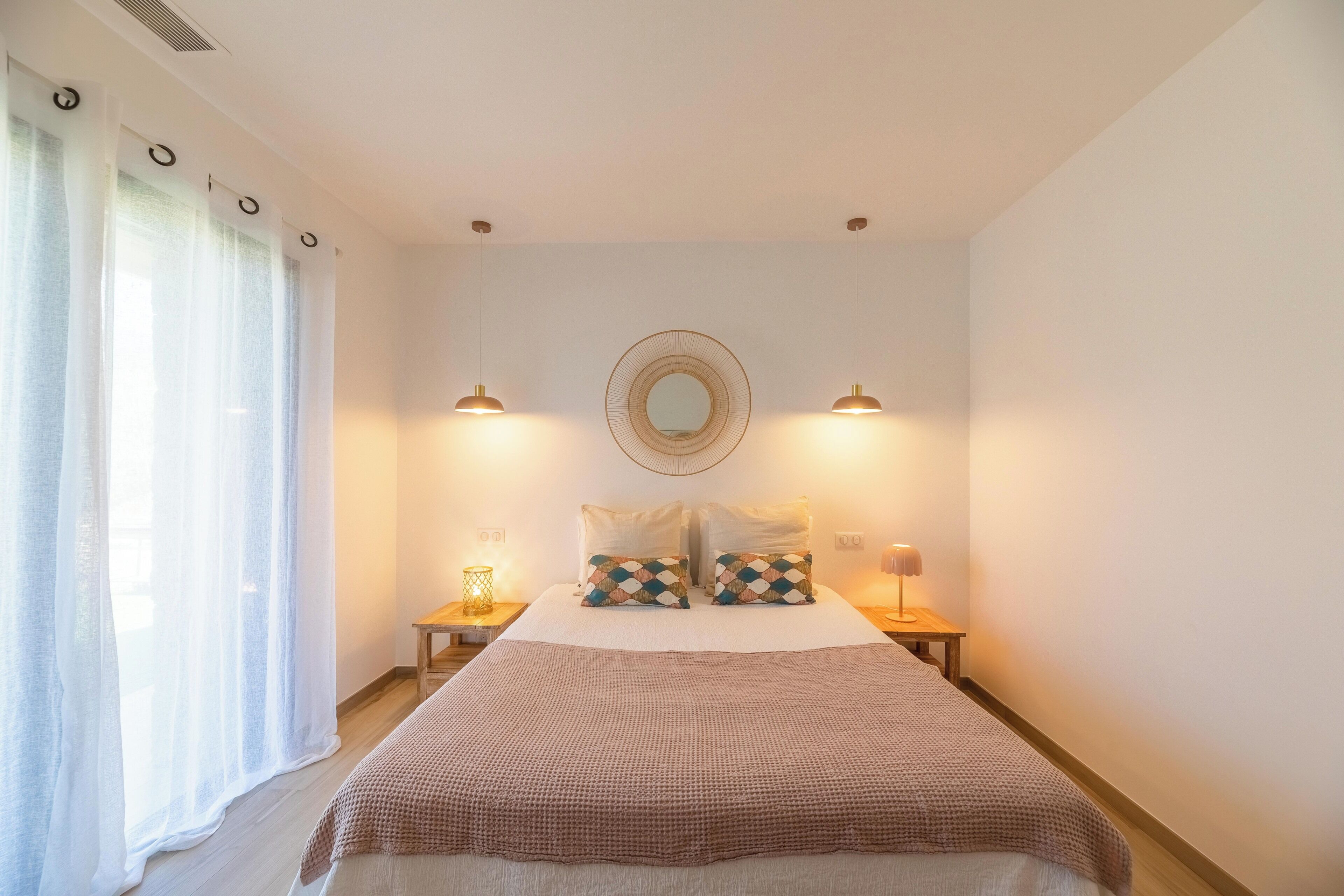 Une chambre à coucher avec un lit, deux lampes de chevet, un miroir rond et une fenêtre avec des rideaux.
