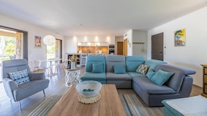 A modern living room with a grey sectional sofa, a small armchair, and a coffee table. There is a kitchen in the background with white cabinets and a dining area with clear chairs. - Villa Lubama 1, Style Bergerie, Entre mer et Montagne Avec Piscine (San-Gavino-Di-Carbini)
