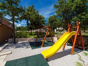 Children can play safely on the playground with a slide and swing set on a gravel surface surrounded by trees and a building.