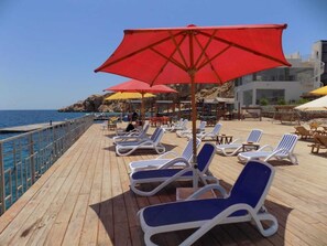 A wooden deck with sun loungers and red umbrellas, overlooking the sea.