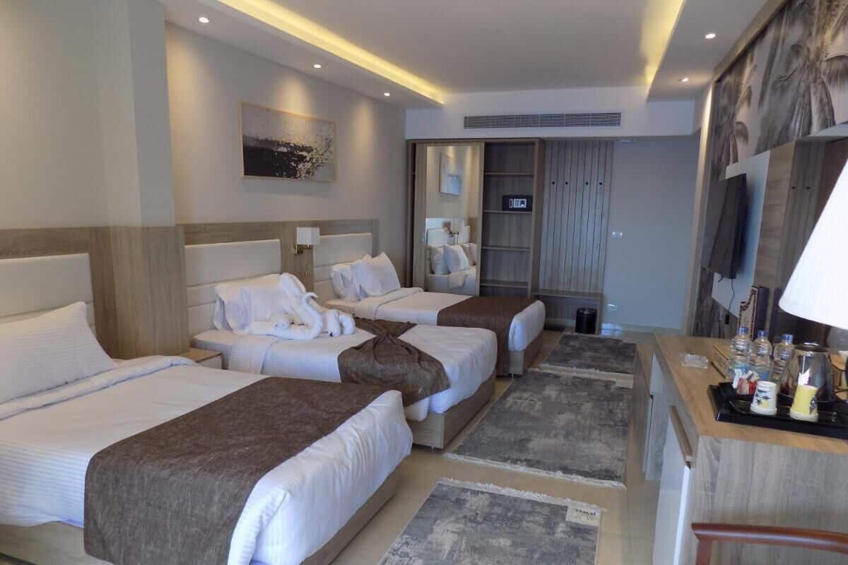 A hotel room with three beds, a desk with a lamp, and a large window.