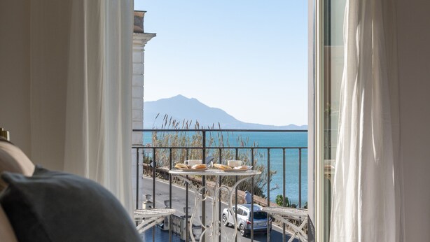 A hotel room with a bed, a view of a coastal area, and a mountain in the distance. - Naro Suites and Rooms (Bacoli)