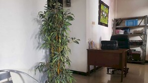 A room with a tall potted plant, a metal chair, a desk with a computer, and a shelving unit with various items. - OYO Life 93202 Pentagon Homestay Syariah (Surabaya)