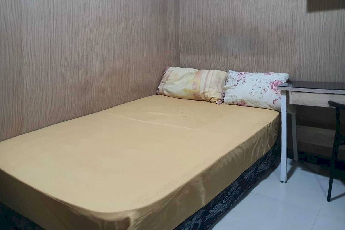 A small, single-bed room with a plain beige mattress, a floral pillow, and a white table with a chair.