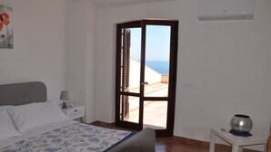 Une chambre à coucher avec un lit, une table de chevet, une petite table ornée d’un vase, et une vue sur la mer par une porte ouverte.