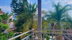 Un balcon doté d’une balustrade en métal, donnant sur un jardin planté de palmiers et sur un bâtiment visible au loin.