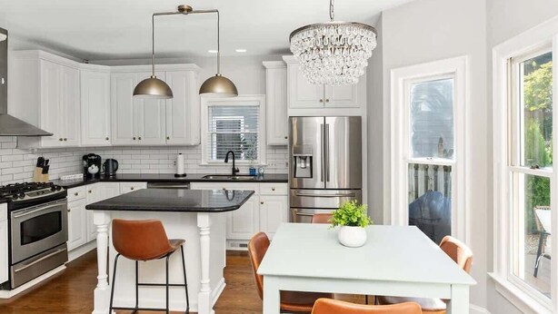 A modern kitchen with white cabinetry, stainless steel appliances, a central island with bar stools, pendant lighting, and a chandelier.