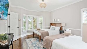 A bedroom with two beds, a desk, a chair, a window with blinds, a ceiling light, and a painting on the wall.