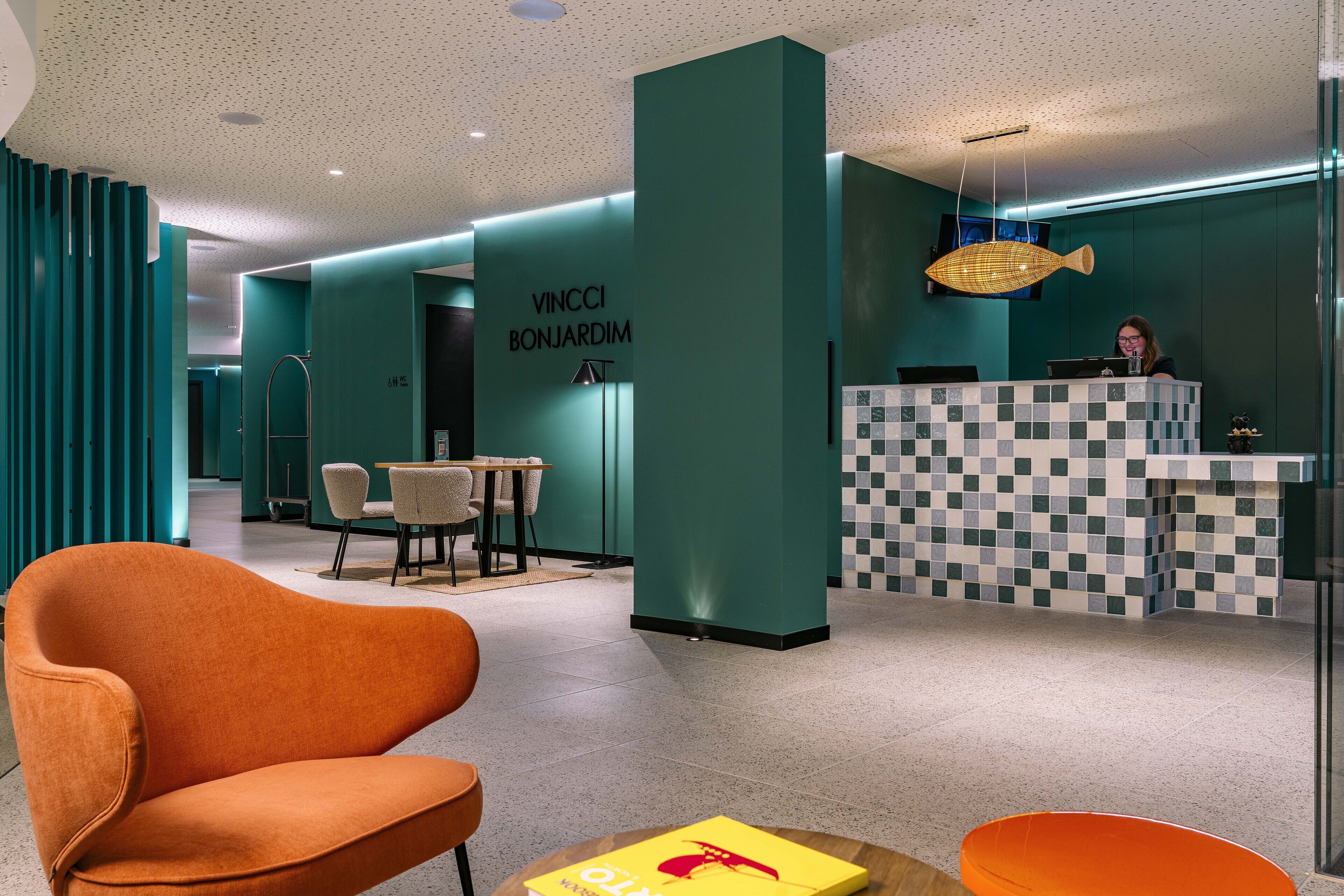 Un vestíbulo moderno con paredes azul verdoso, un mostrador de recepción a cuadros y un sillón naranja grande.