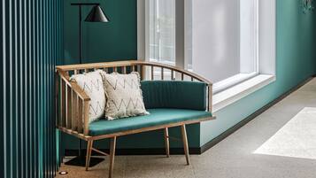 A corner room with a teal wall, a wooden bench with cushions, a floor lamp, and a large window.