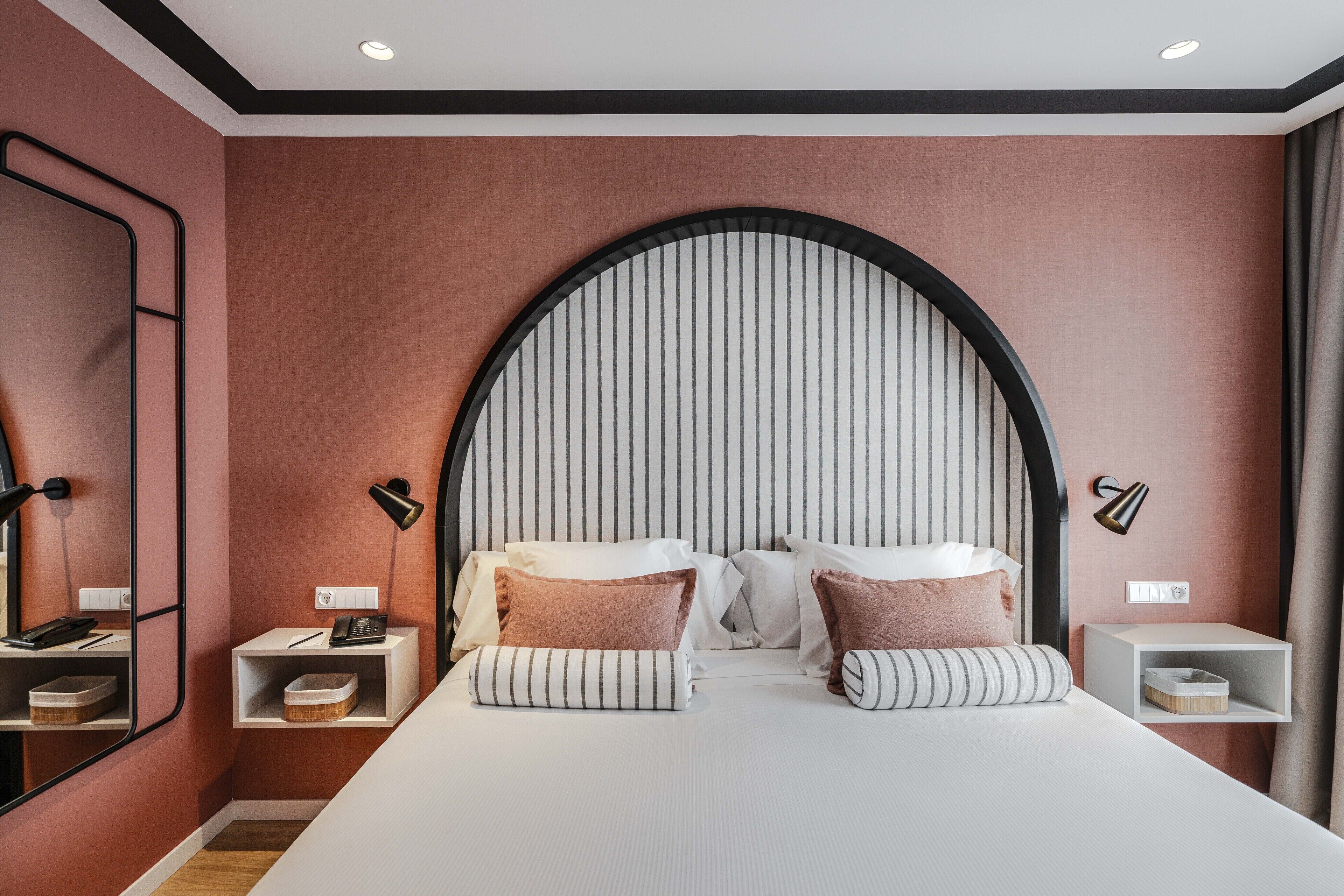 A modern hotel room with a large bed, two bedside tables, a mirror, and a unique arched headboard.
