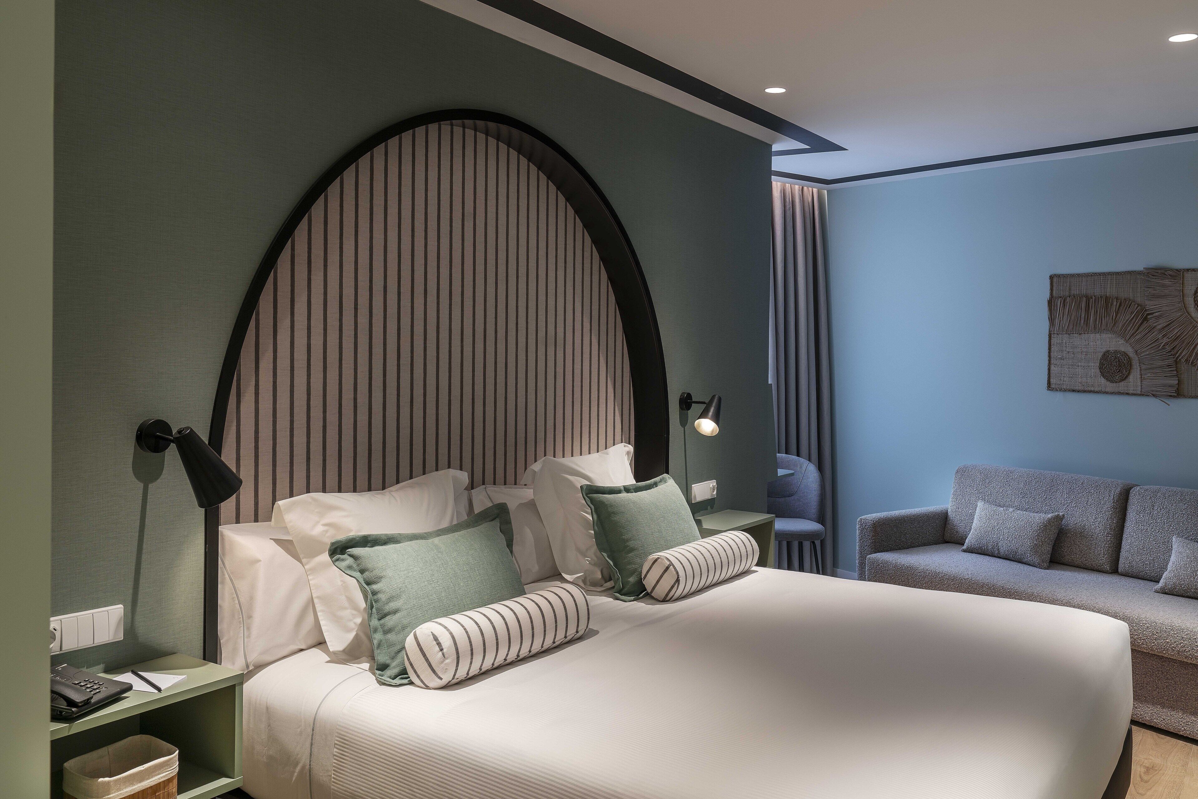A modern bedroom with a large bed, a nightstand, a bedside table, a sofa, and a wall-mounted lamp.