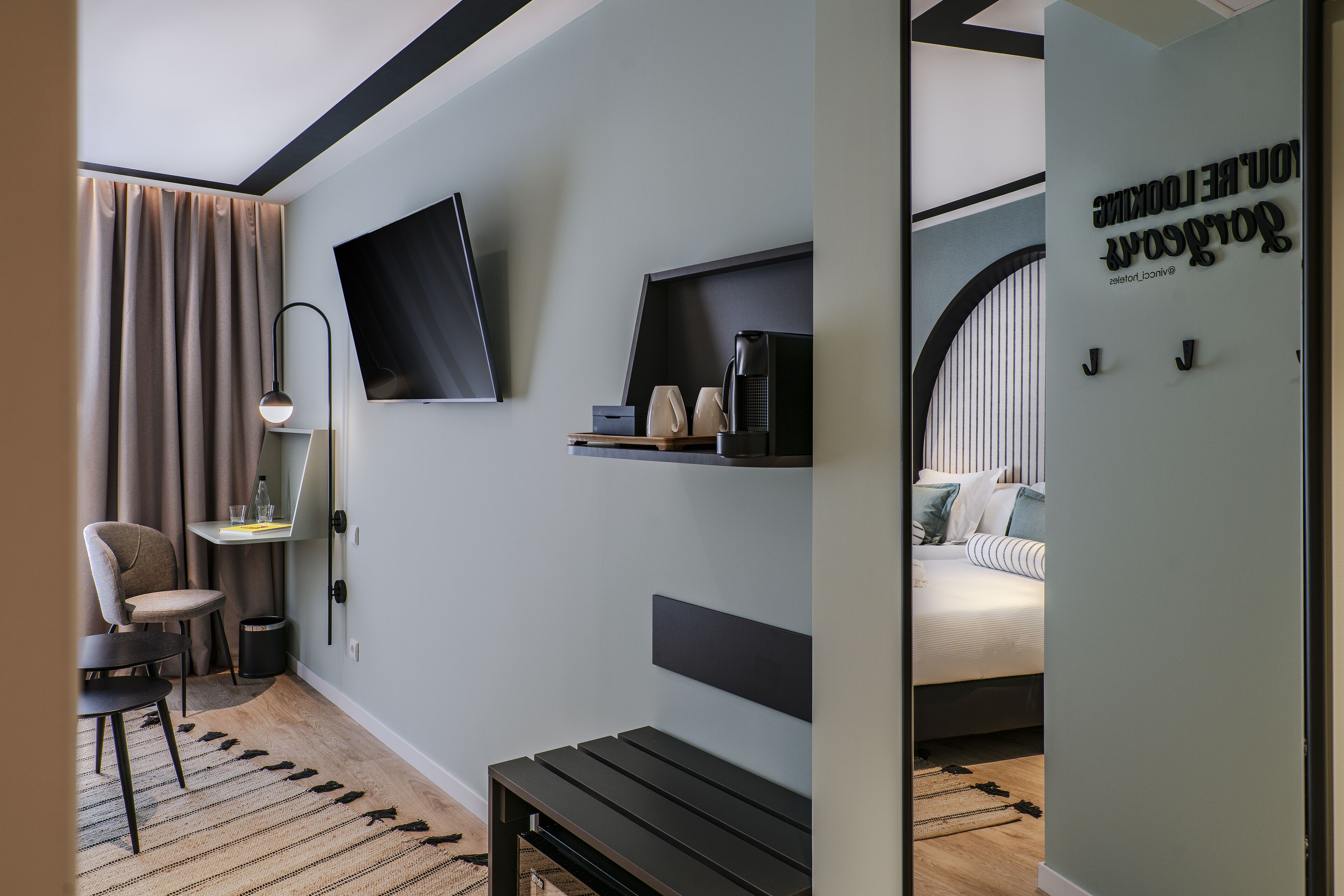 A modern hotel room with a bed, a desk with a flat-screen TV, a coffee machine, and a chair.